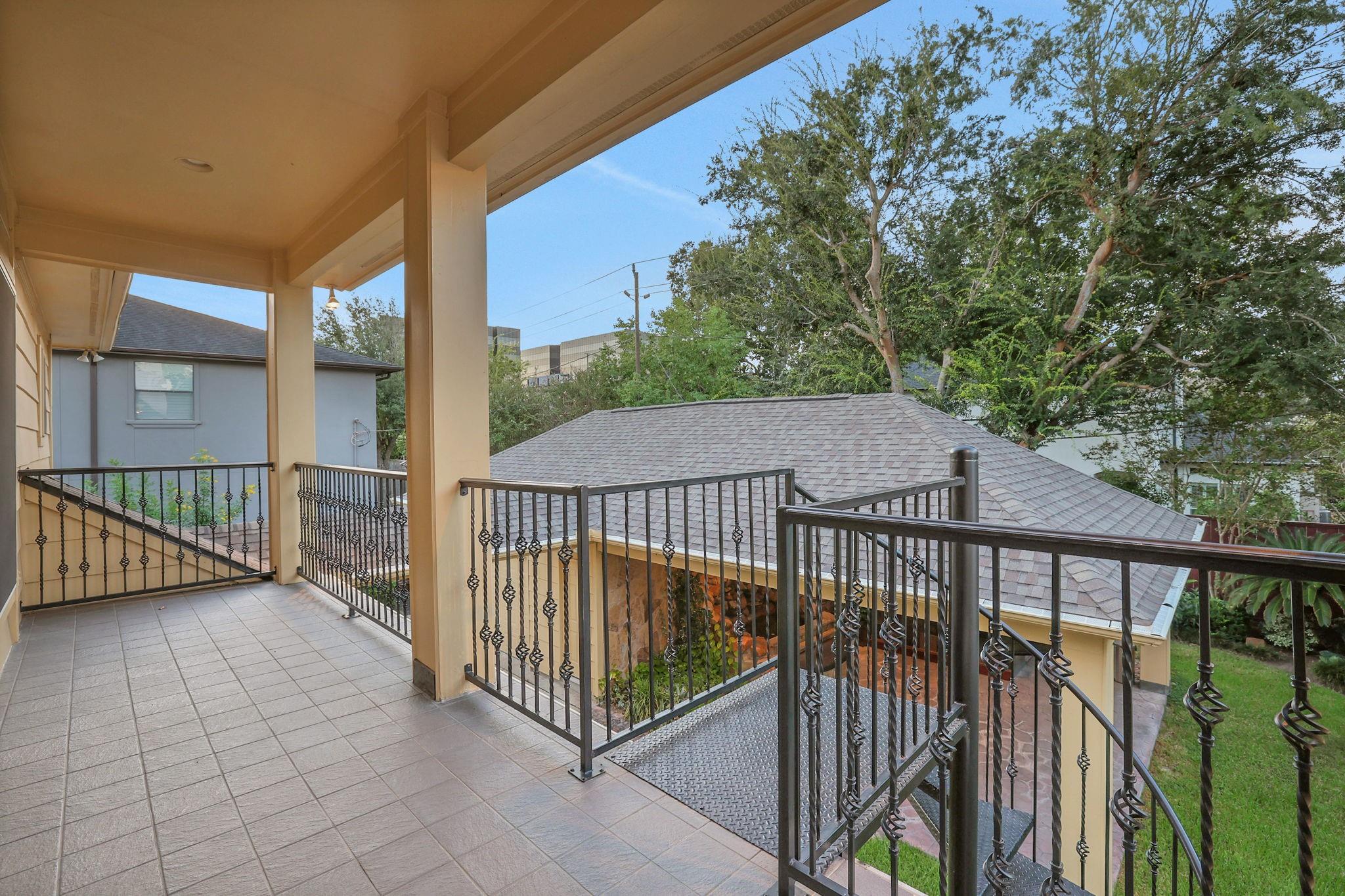 Covered tiled balcony with ornate railings and a metal staircase overlooking trees and neighboring rooftops.