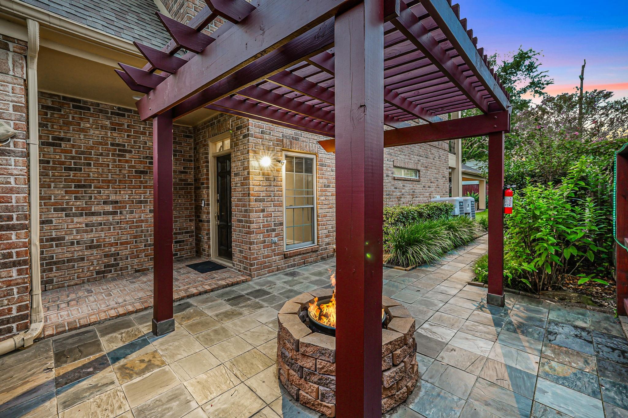 Stone patio with pergola and burning fire pit beside a brick home
