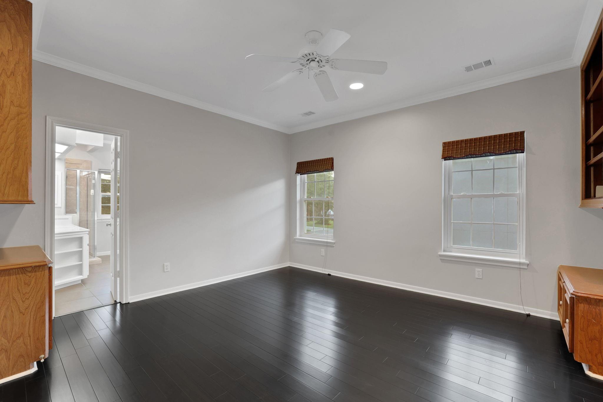 Empty bedroom with dark hardwood floors, two windows, a ceiling fan, and an open doorway to the bathroom.