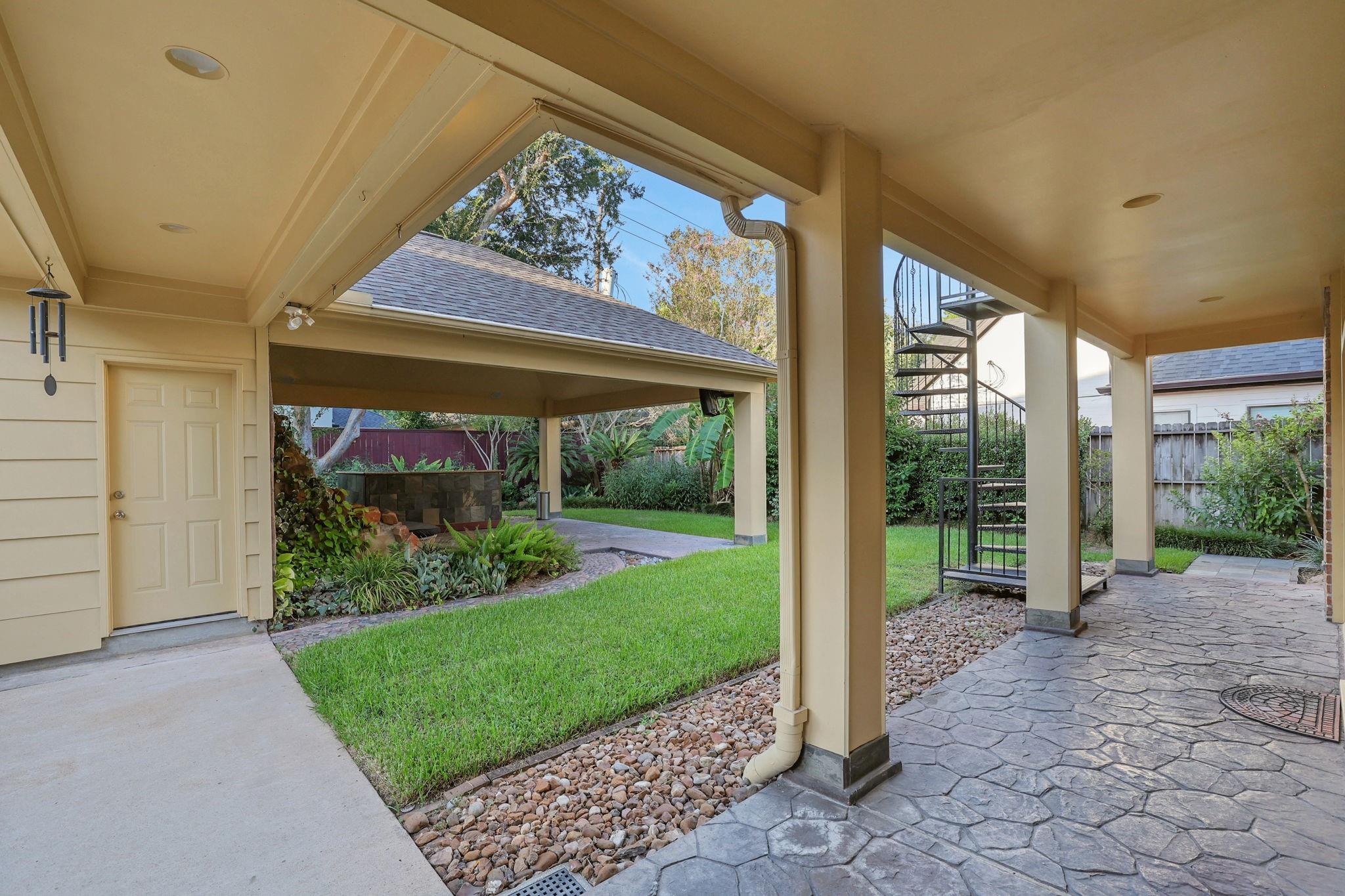 Covered backyard patio with stamped-stone flooring, a green lawn, landscaped garden, and black spiral staircase.