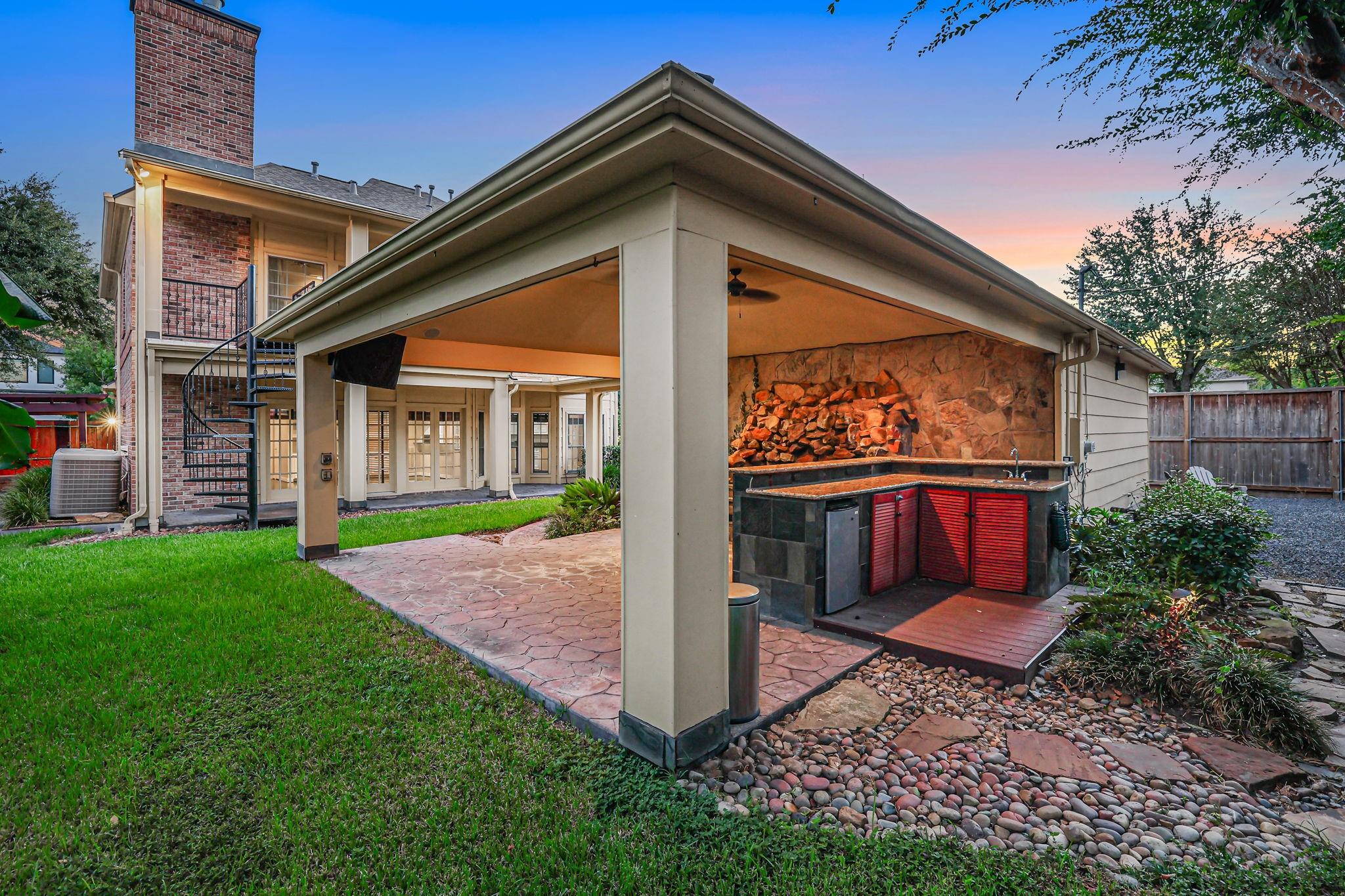 Covered outdoor kitchen with stone fireplace and red cabinets beside a brick home.