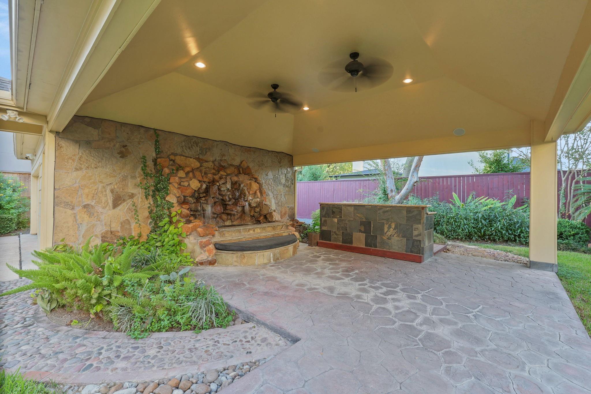 Covered stone patio with a rock waterfall, curved basin, ceiling fans, and tiled counter.