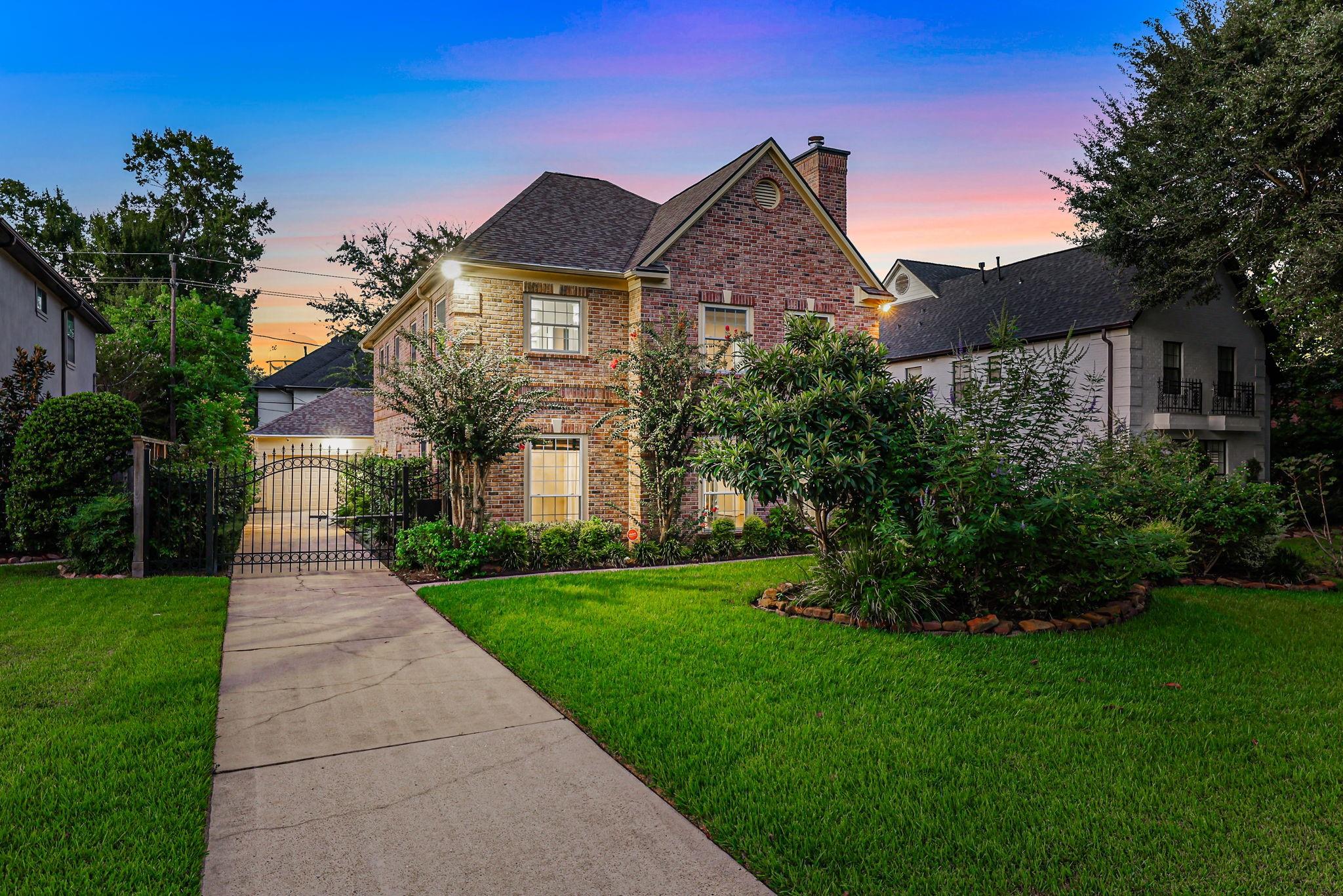 Two-story brick house at dusk, with a gated driveway, broad lawn, and mature landscaping.