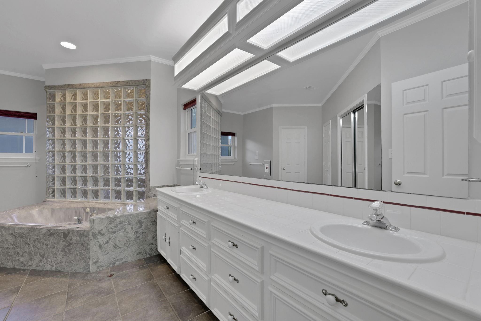 Bathroom with a long double vanity, skylights, glass-block partition, and corner tub.