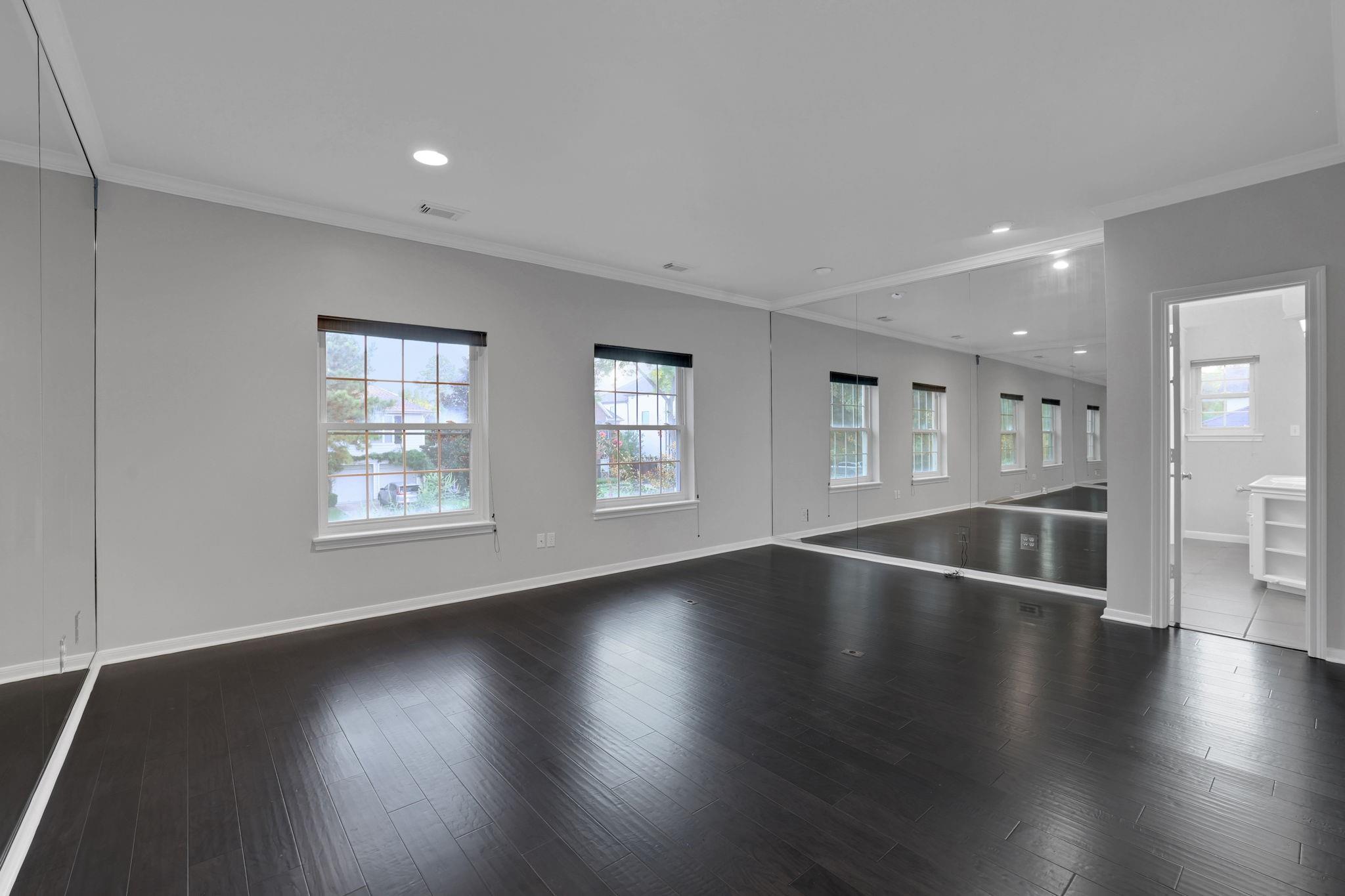 Vacant room with a mirrored wall, dark wood flooring, and windows beside a bathroom doorway.