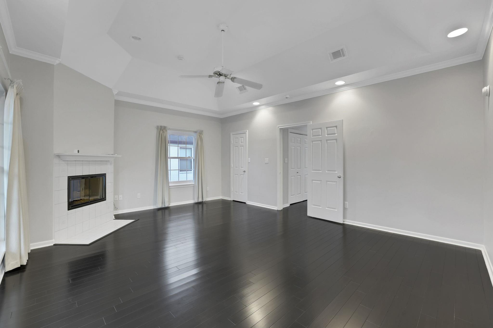Empty living room with dark hardwood floors, a white tiled fireplace, and a tray ceiling fan.