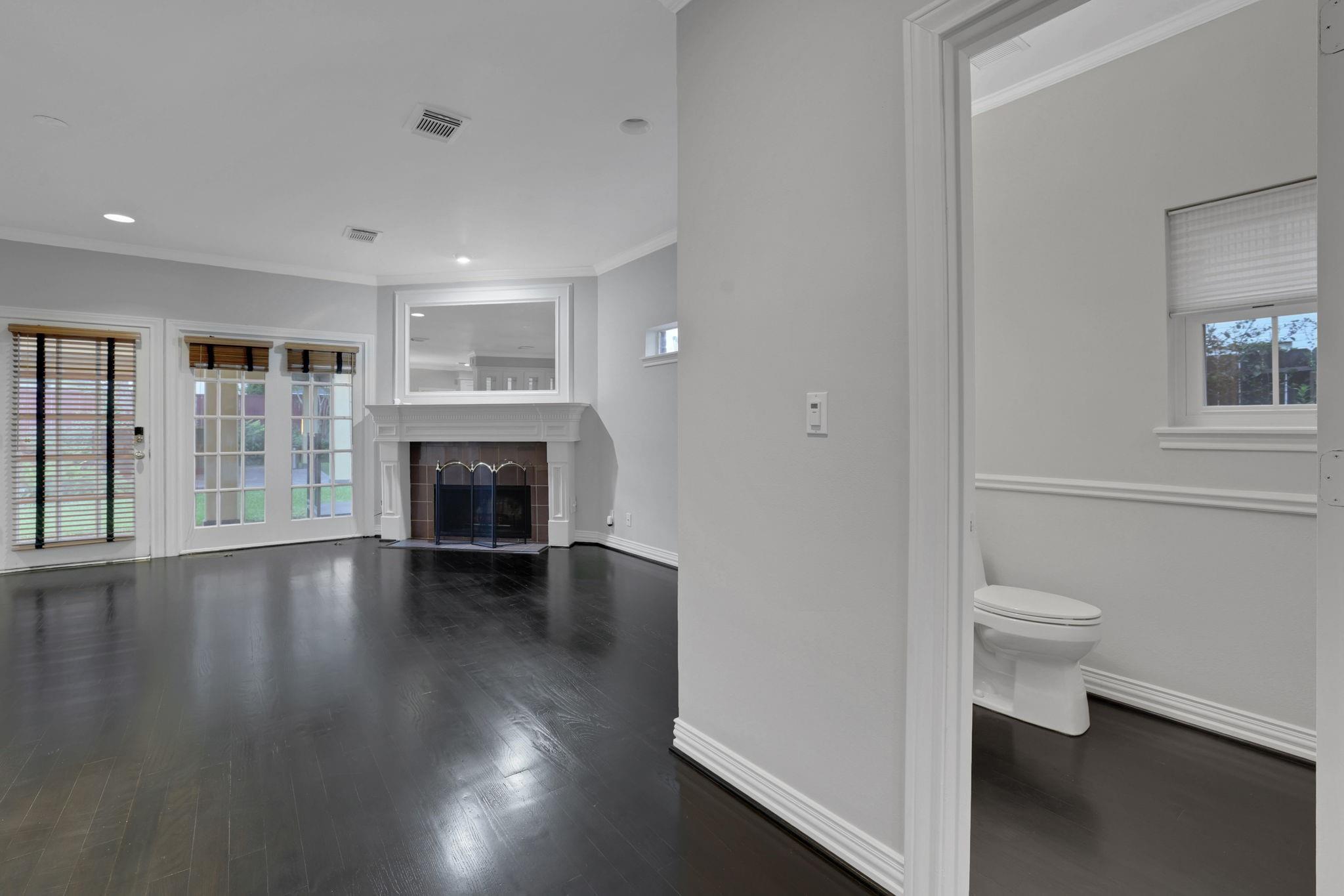 Vacant living room with dark hardwood floors, white fireplace, and French doors; adjoining bathroom visible.