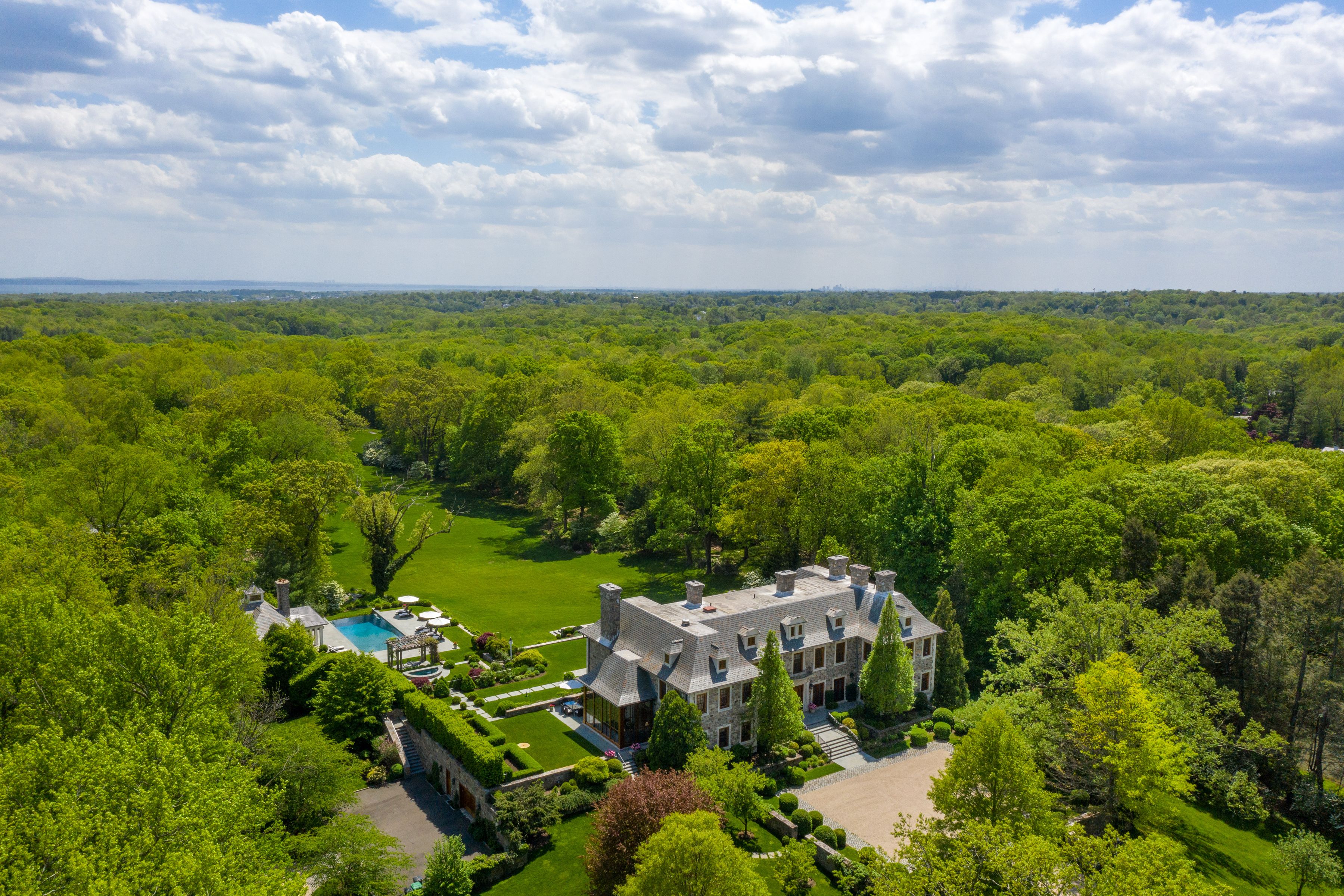Stone estate amid dense woodland, with a swimming pool, broad lawn, and landscaped grounds.