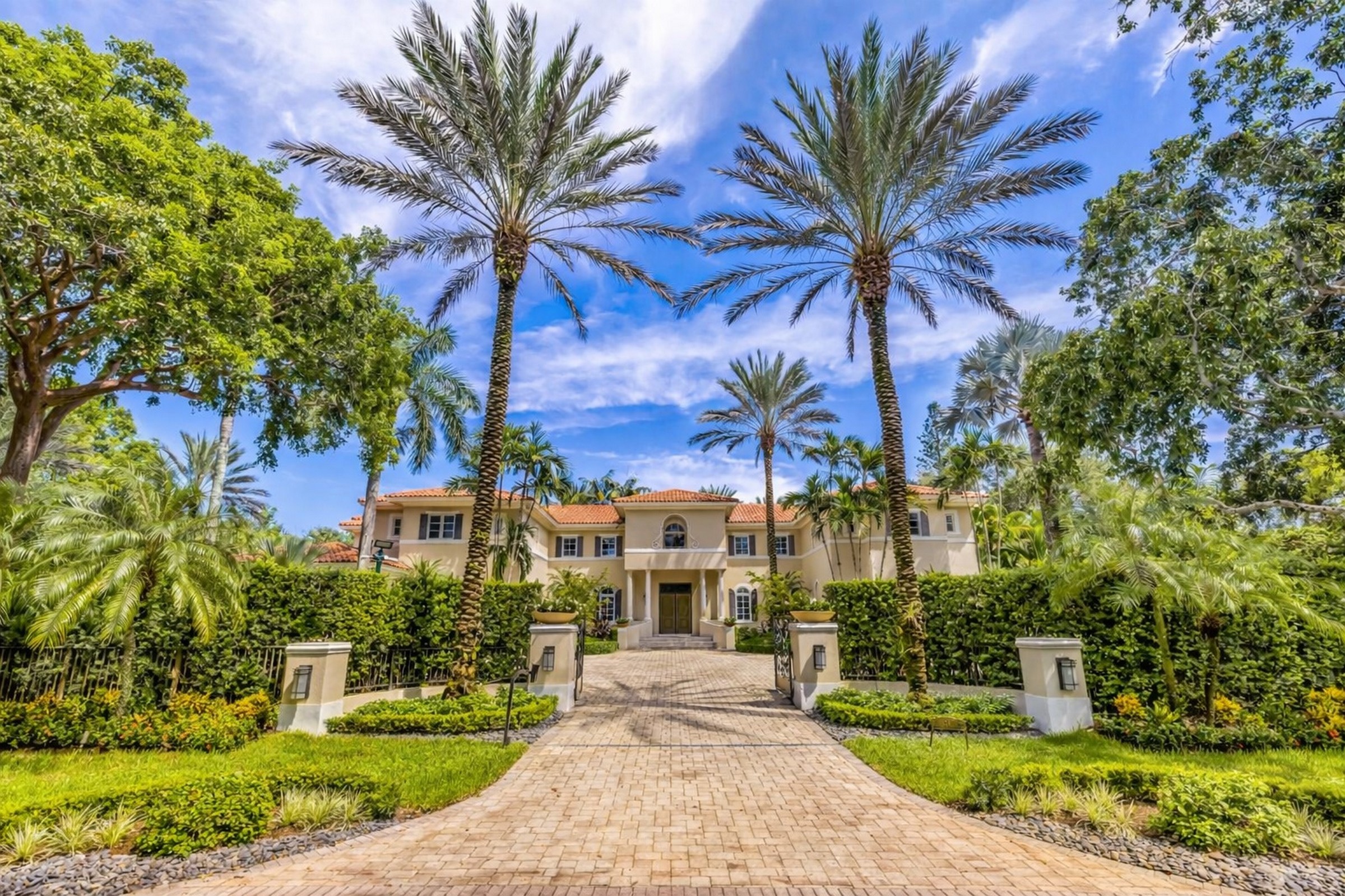 Large cream-colored home framed by tall palm trees and tropical landscaping at the end of a brick driveway.