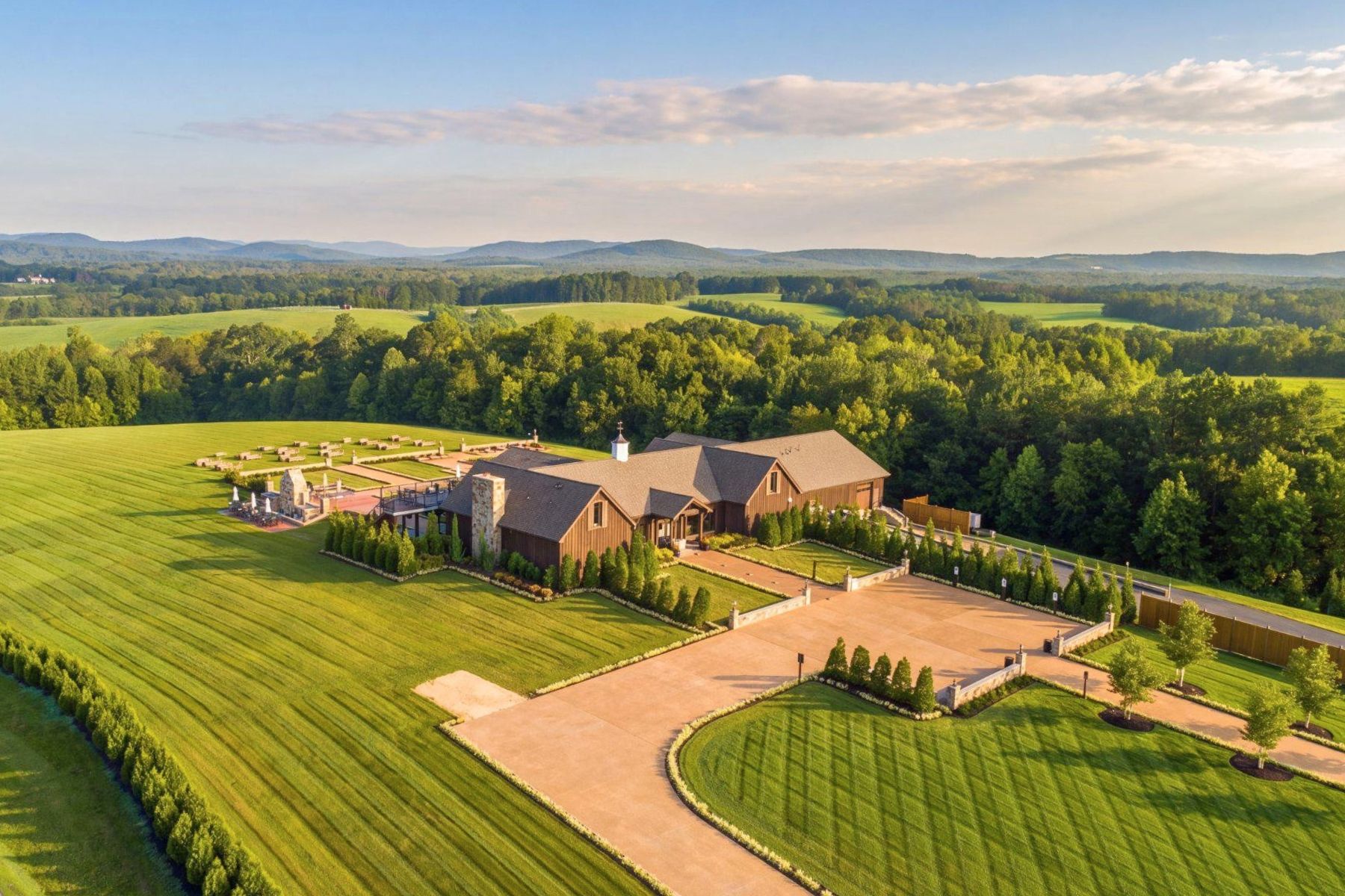 Aerial view of a country home with broad lawns and a long driveway, backed by fields and wooded hills.