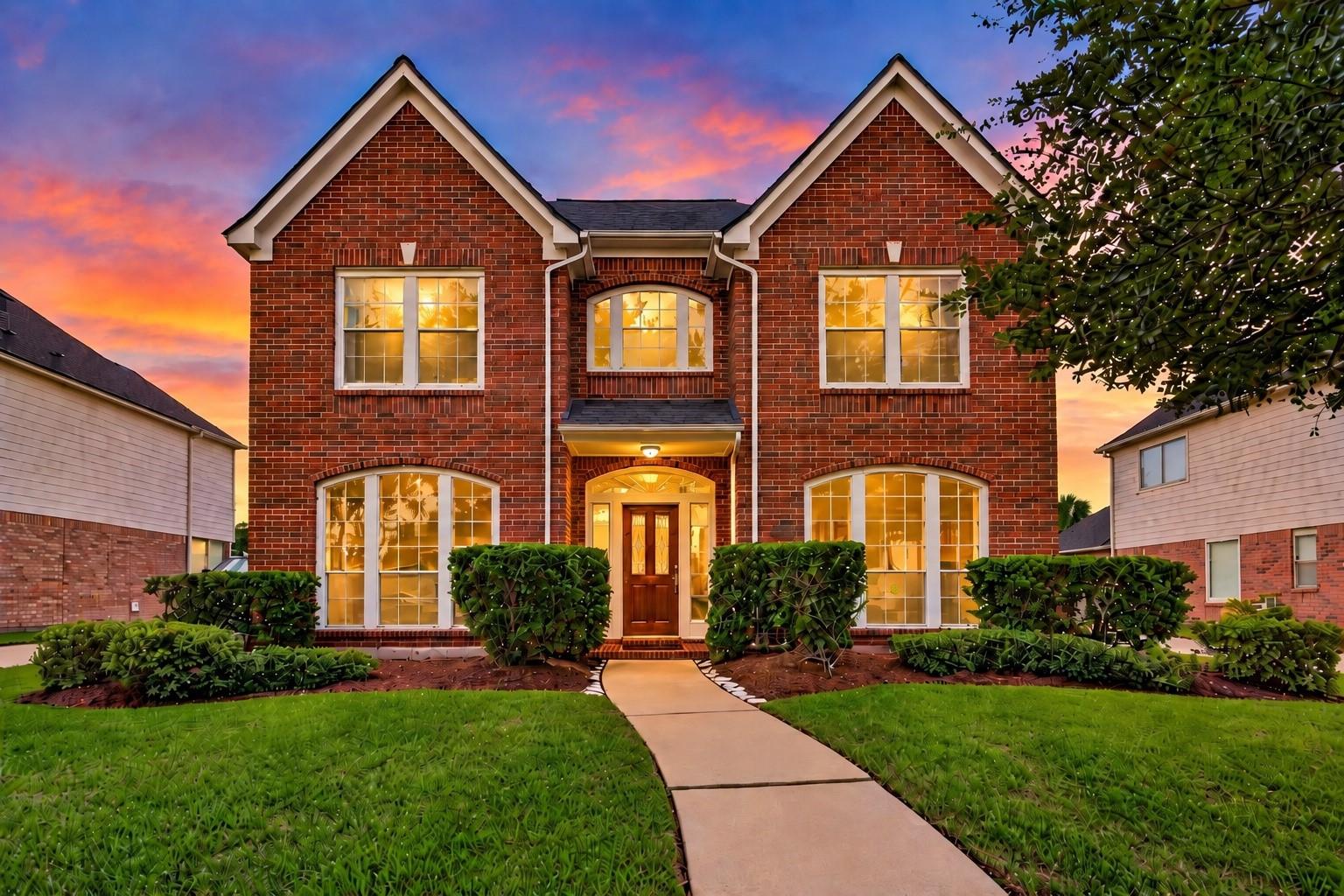 Two-story brick house with glowing windows, trimmed hedges, and a curved front walkway at sunset.