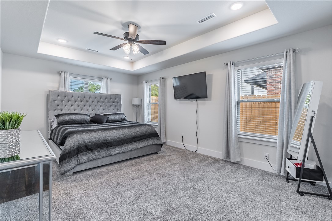 Bedroom with gray tufted bed, tray ceiling, wall-mounted TV, and carpeted floor.