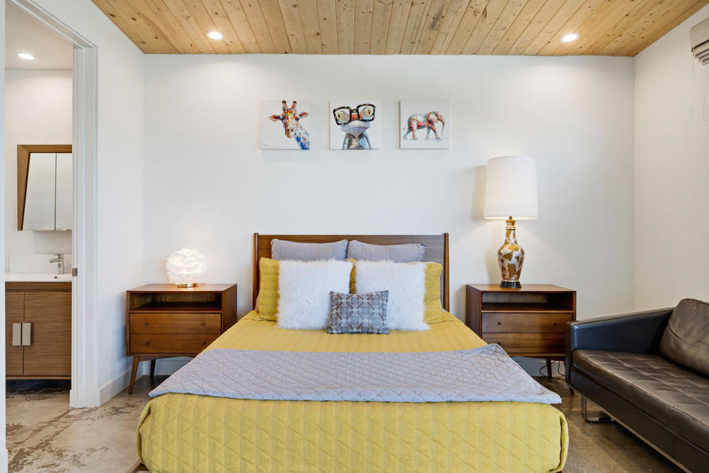 Staged bedroom with yellow bedding, a wood-plank ceiling, colorful animal art, and an adjoining bathroom.