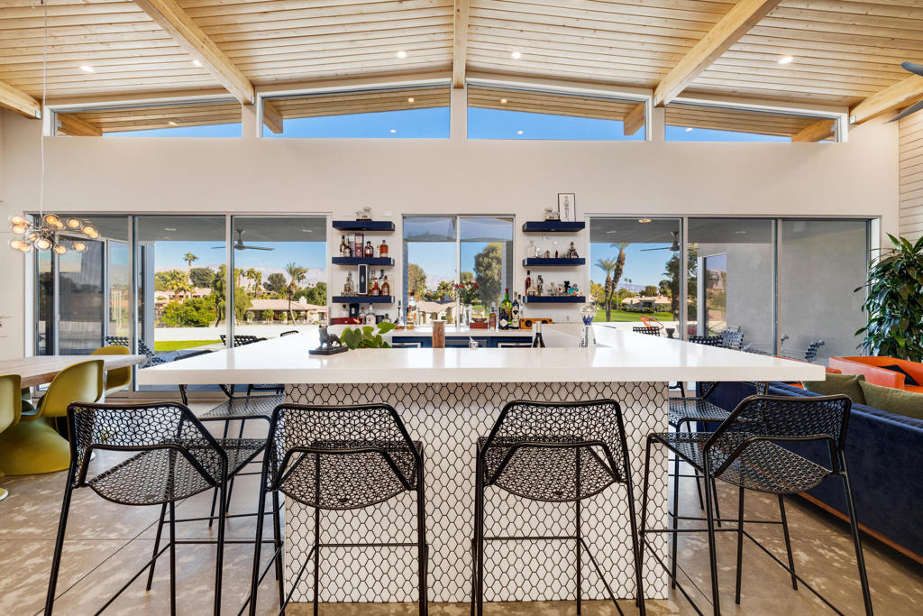 Kitchen with a long white island, woven bar stools, and a high wood-plank ceiling.