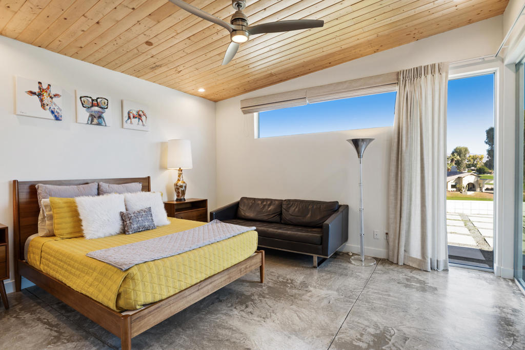 Bedroom with a yellow-quilted bed, dark sofa, wood-plank ceiling, and glass doors to a patio.