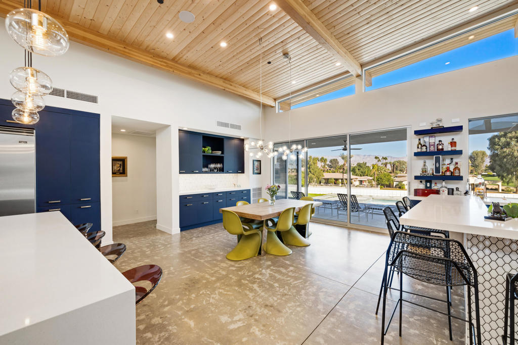 Modern kitchen and dining area with dark blue cabinets, a vaulted wood ceiling, and pool views through glass doors.