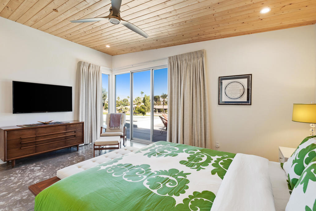 Bedroom with a green patterned bedspread, wood-plank ceiling, and sliding glass doors to a sunny patio.