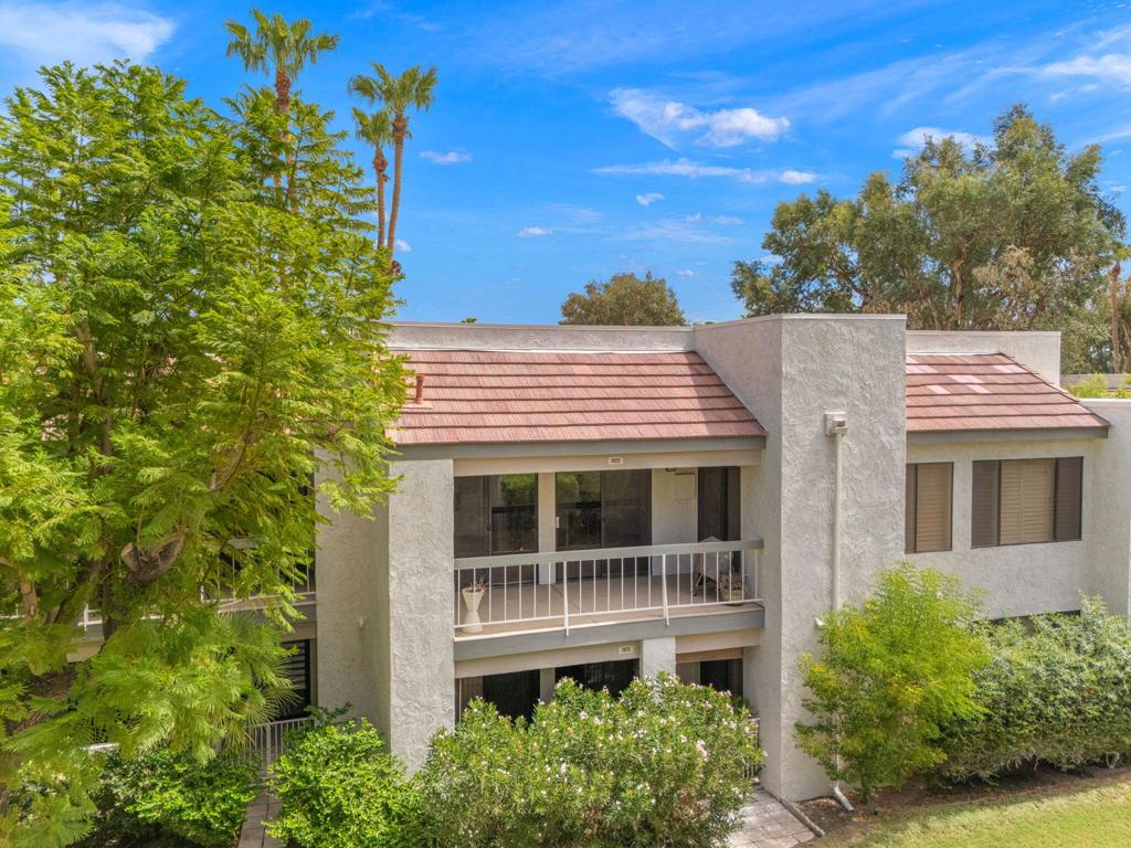 Two-story residence with a red tile roof and balcony, framed by mature trees and palms.