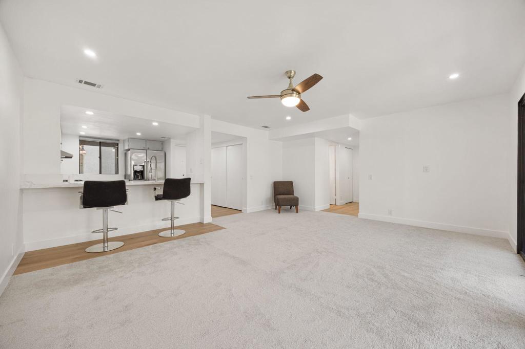 Open living room with a carpeted floor, ceiling fan, and kitchen breakfast bar with two stools.