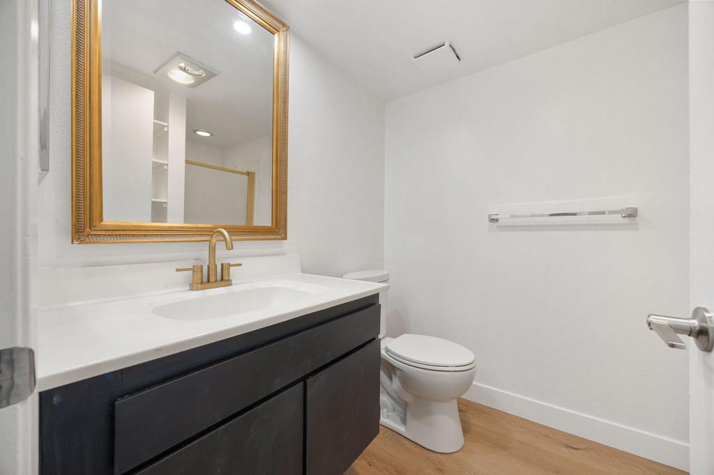 Bathroom with a dark vanity, gold-framed mirror, toilet, and light wood flooring.