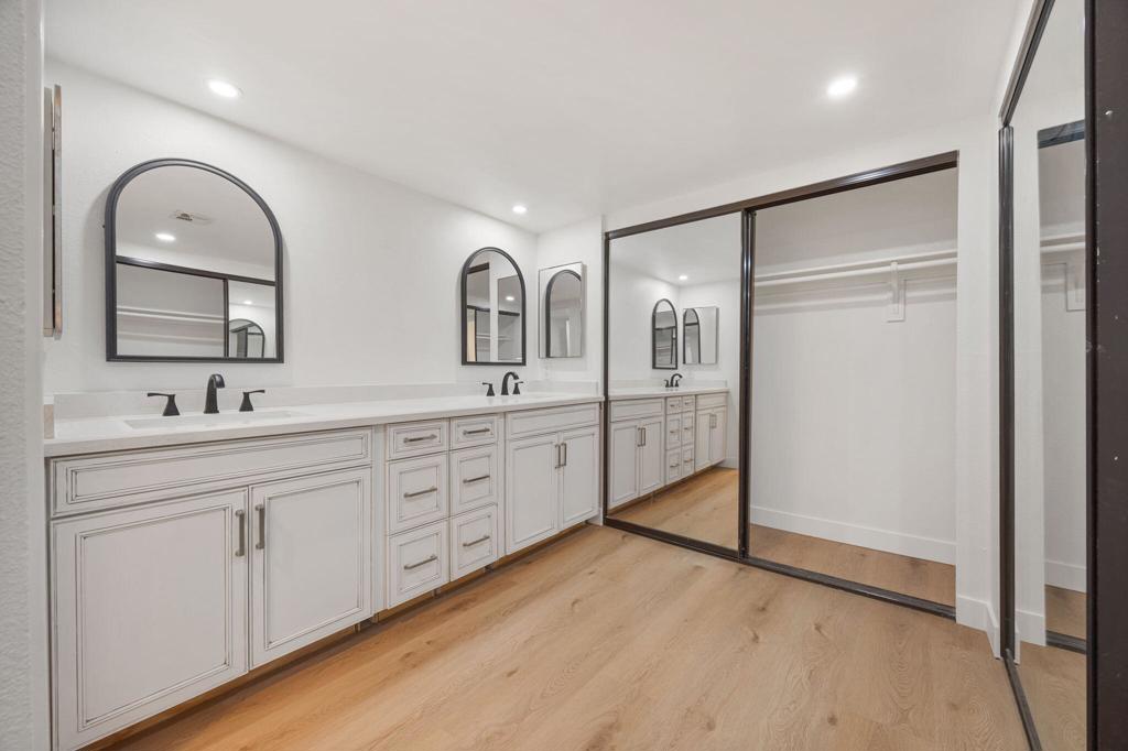 Bathroom with a long white double vanity, arched mirrors, mirrored closet doors, and light wood flooring.
