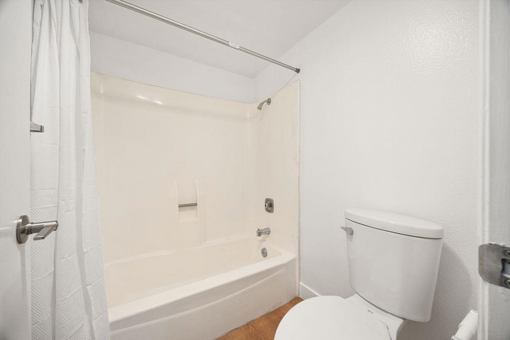 Bathroom with a white tub and shower, curtain, toilet, and wood-look floor.