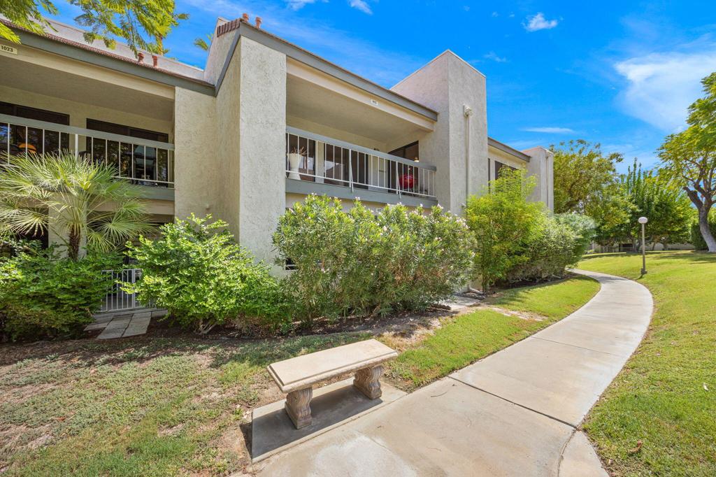 Two-story beige condominium with covered balconies, lush landscaping, and a curved sidewalk.