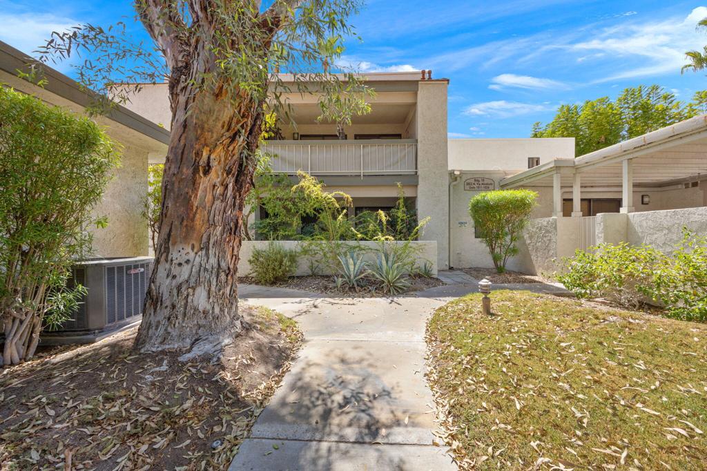 Two-story condominium exterior with a recessed balcony, mature tree, and landscaped walkway.