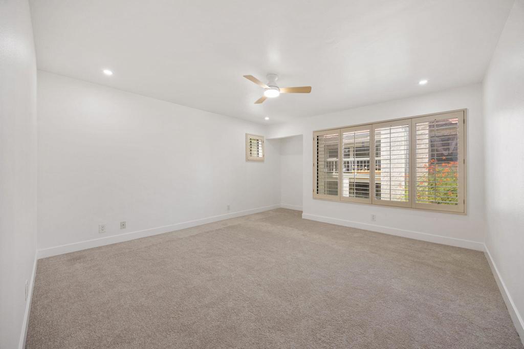 Empty carpeted room with a ceiling fan and a wide shuttered window.