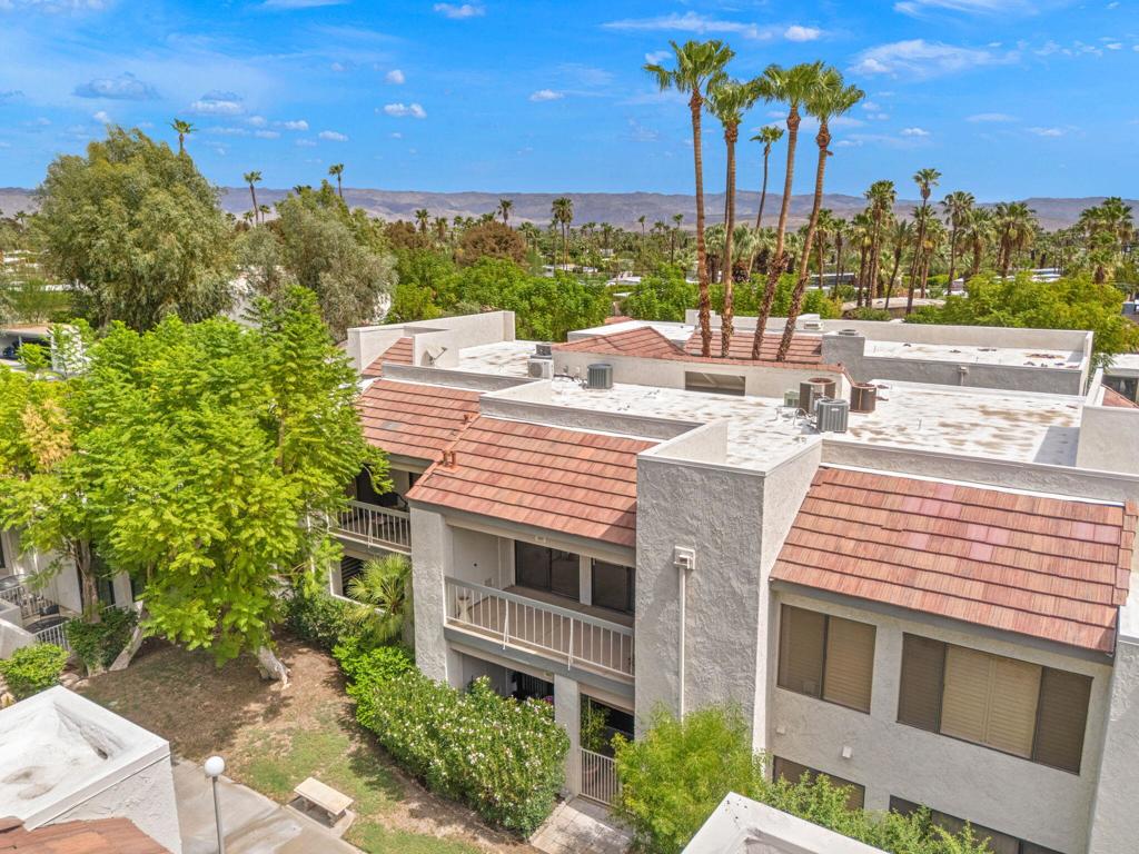 Aerial view of stucco condominium buildings with red tile roofs, palm trees, and distant mountains.