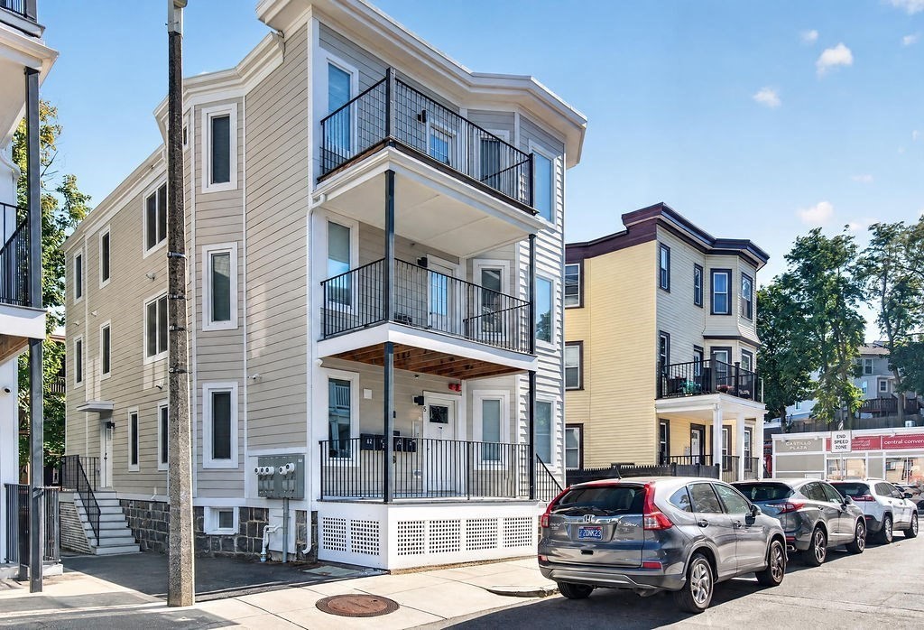 Beige multi-unit building with black balconies and parked cars along the street.