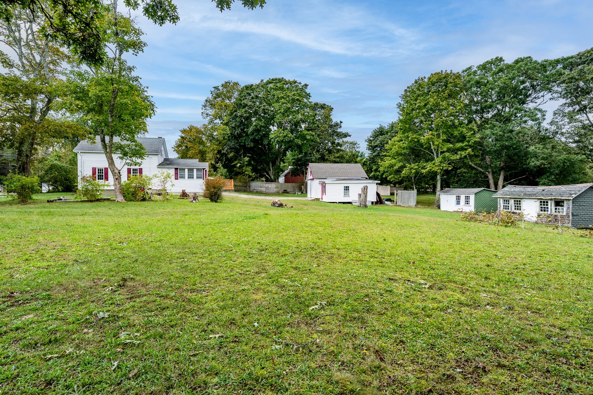 White two-story house and detached outbuildings beyond a wide grassy yard and mature trees.