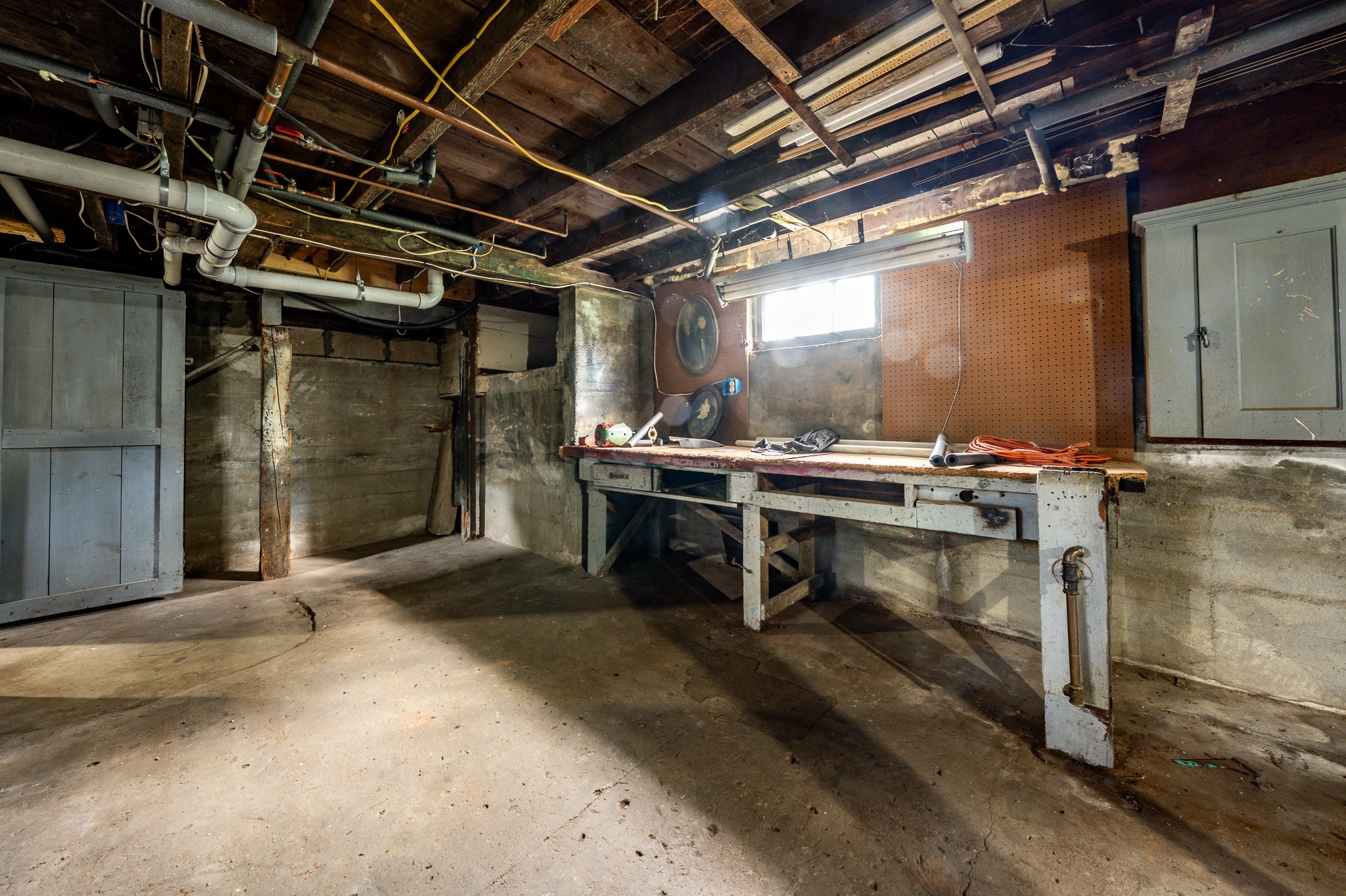 Basement workshop with a long workbench beneath exposed ceiling joists and pipes.