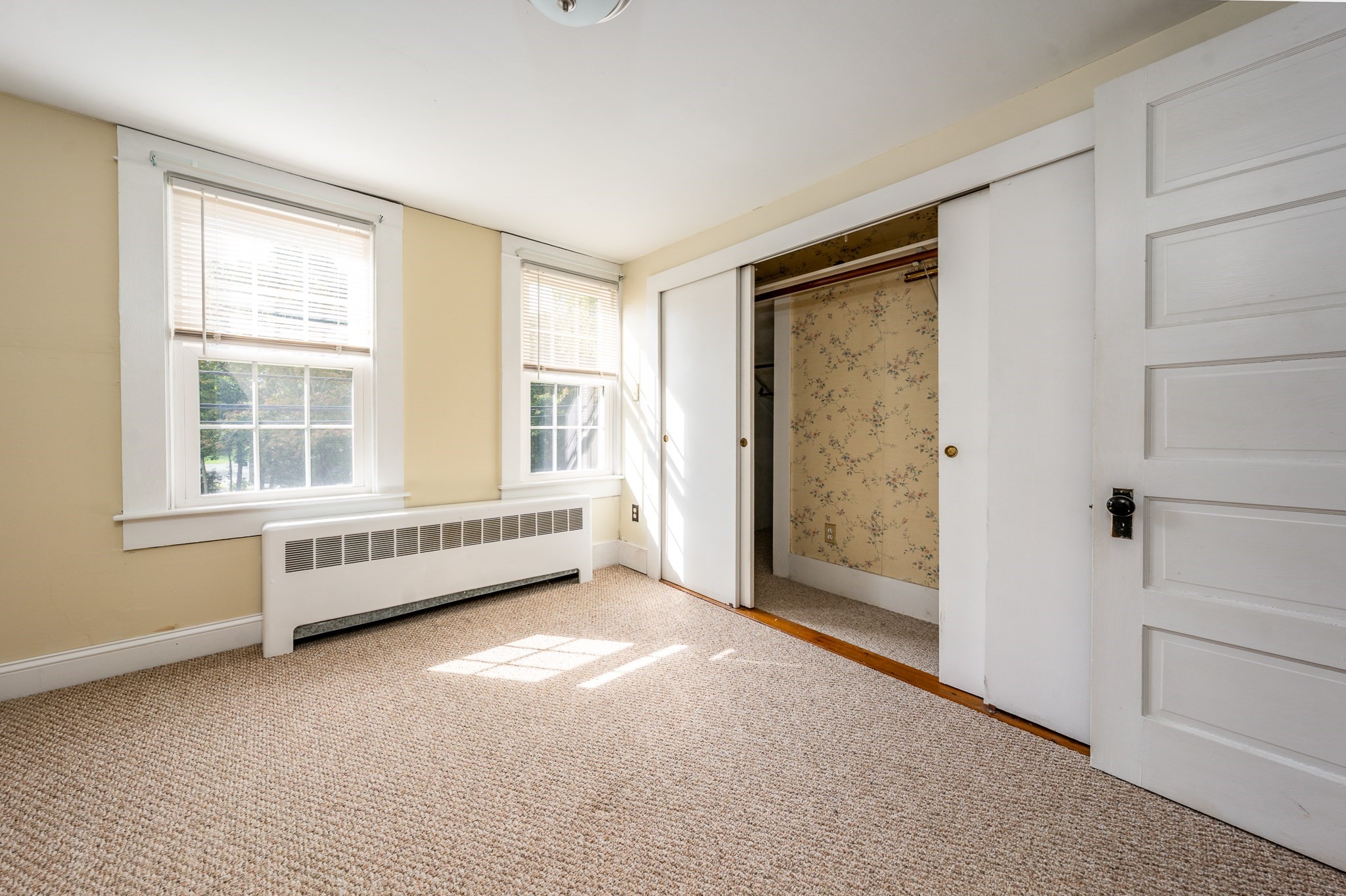 Vacant bedroom with beige carpet, two sunlit windows, a baseboard heater, and an open closet.