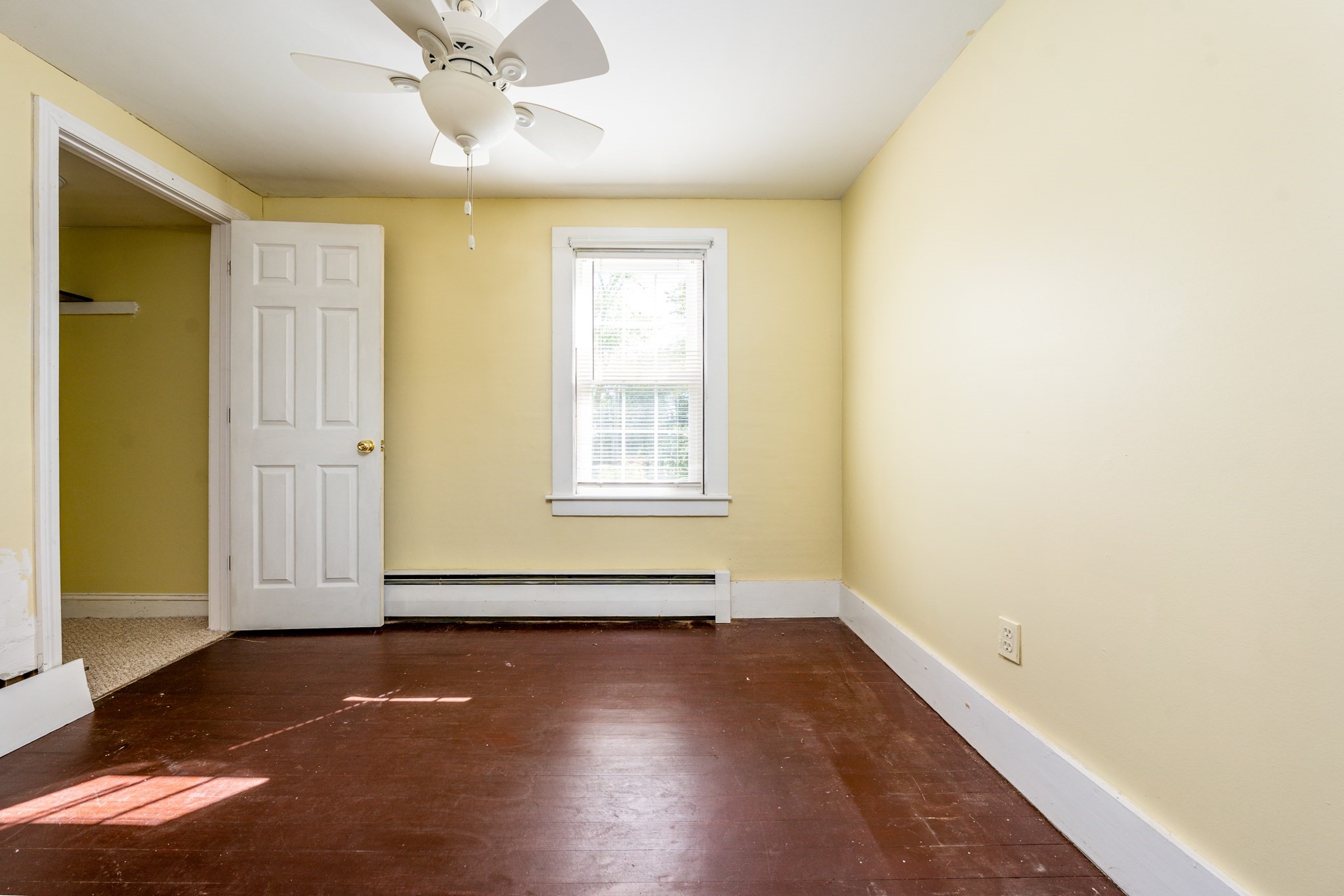 Empty bedroom with yellow walls, dark hardwood floors, a ceiling fan, and a window with blinds.
