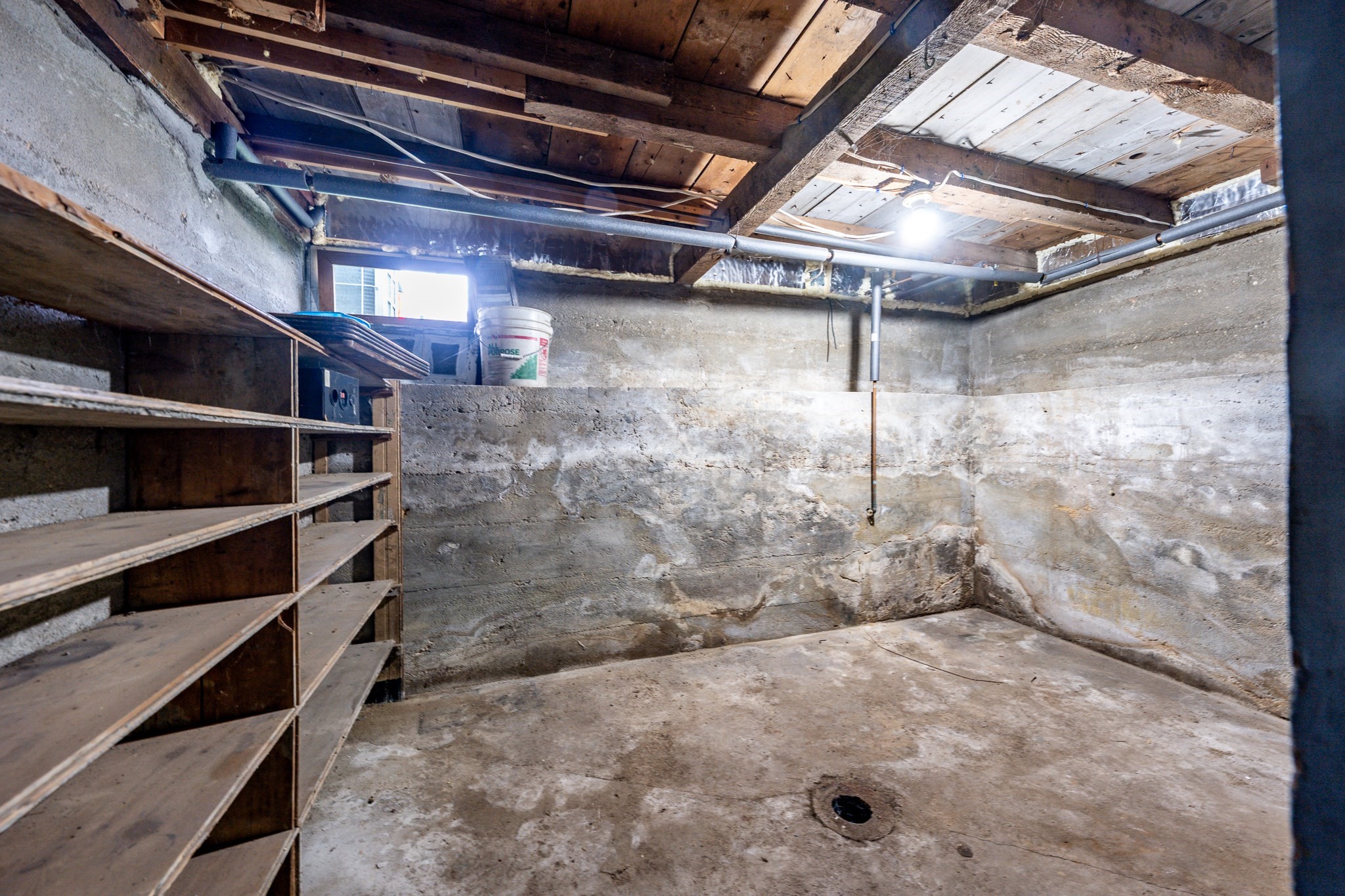 Unfinished basement with concrete walls, exposed ceiling joists, wooden shelving, and a floor drain.