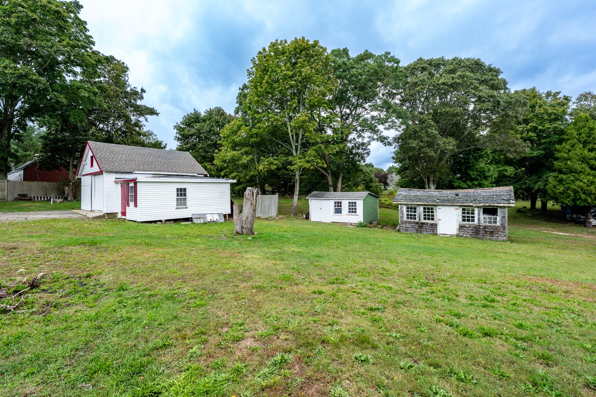 White building with red trim and two detached sheds in a tree-lined grassy yard.
