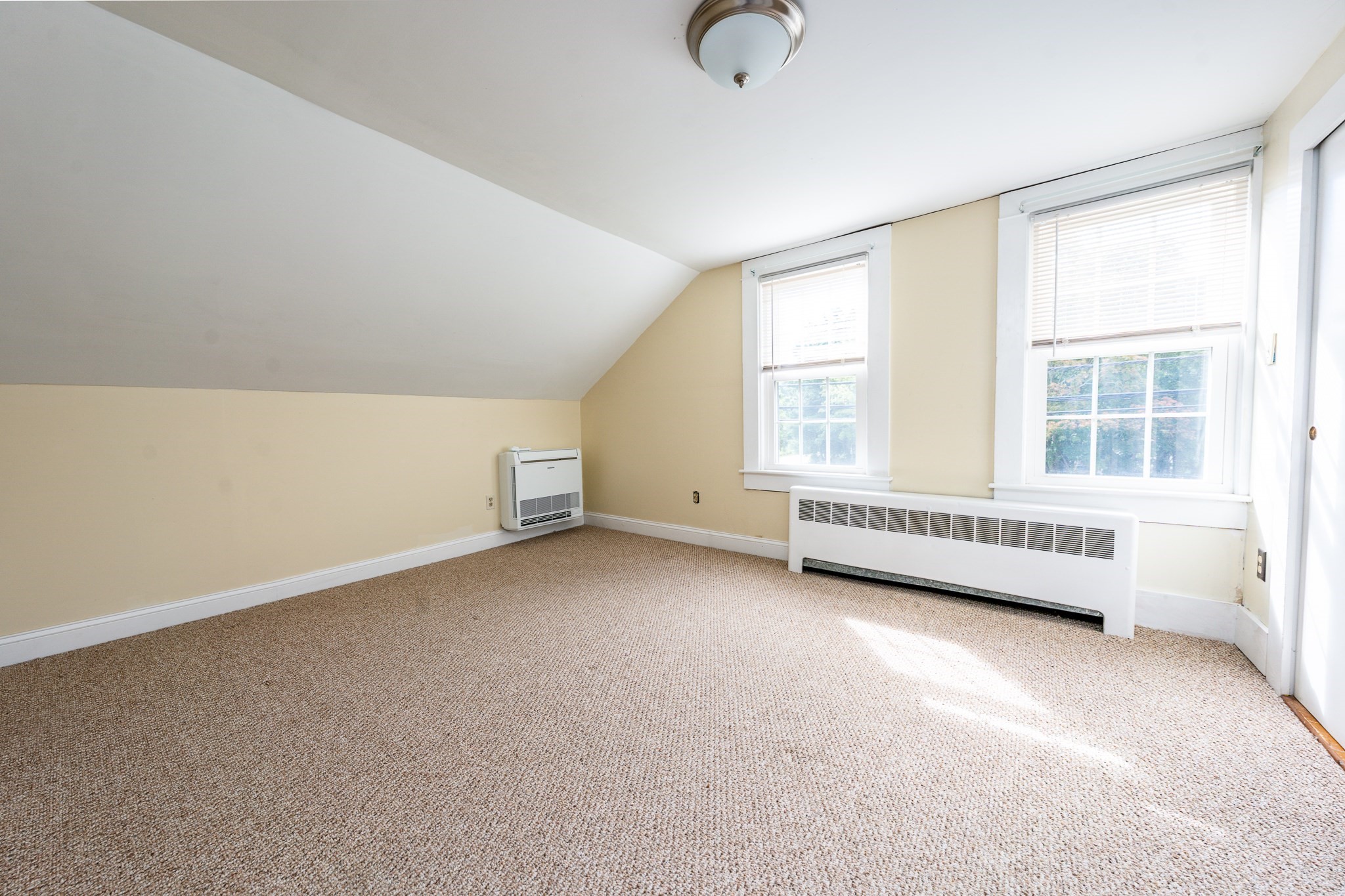 Empty upstairs room with a sloped ceiling, two windows, tan carpet, and baseboard heating.
