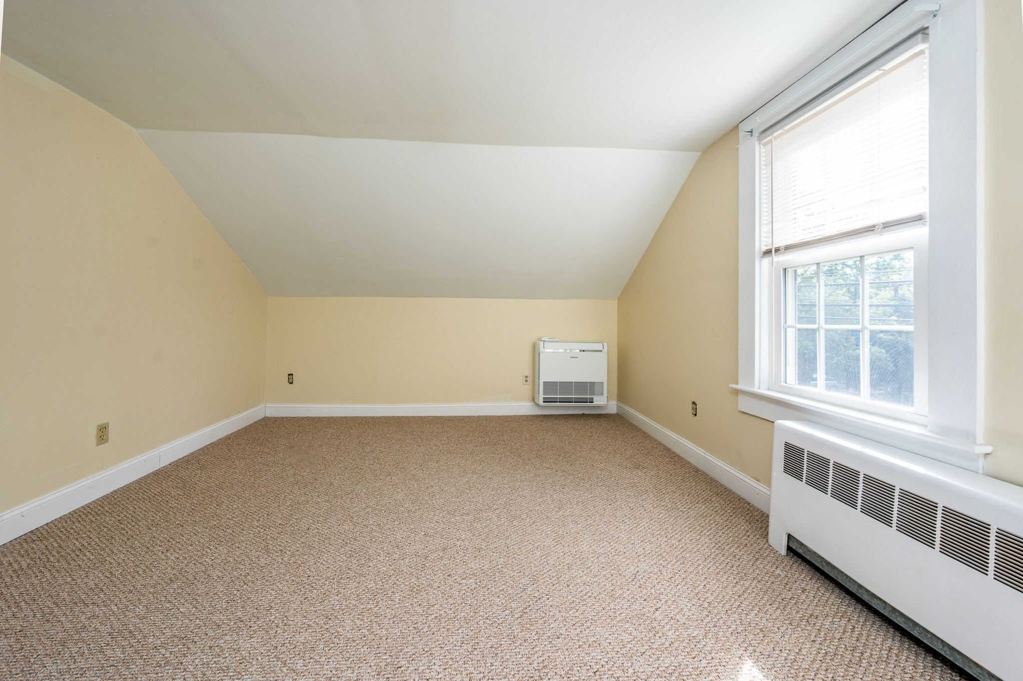 Empty carpeted room with a sloped ceiling, large window, wall-mounted air conditioner, and baseboard heater.
