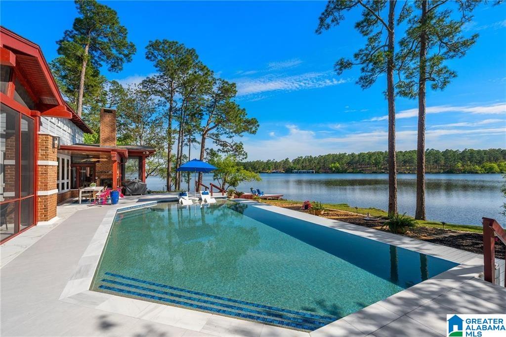 Lakeside swimming pool beside a modern home, with a stone patio and tall pine trees.