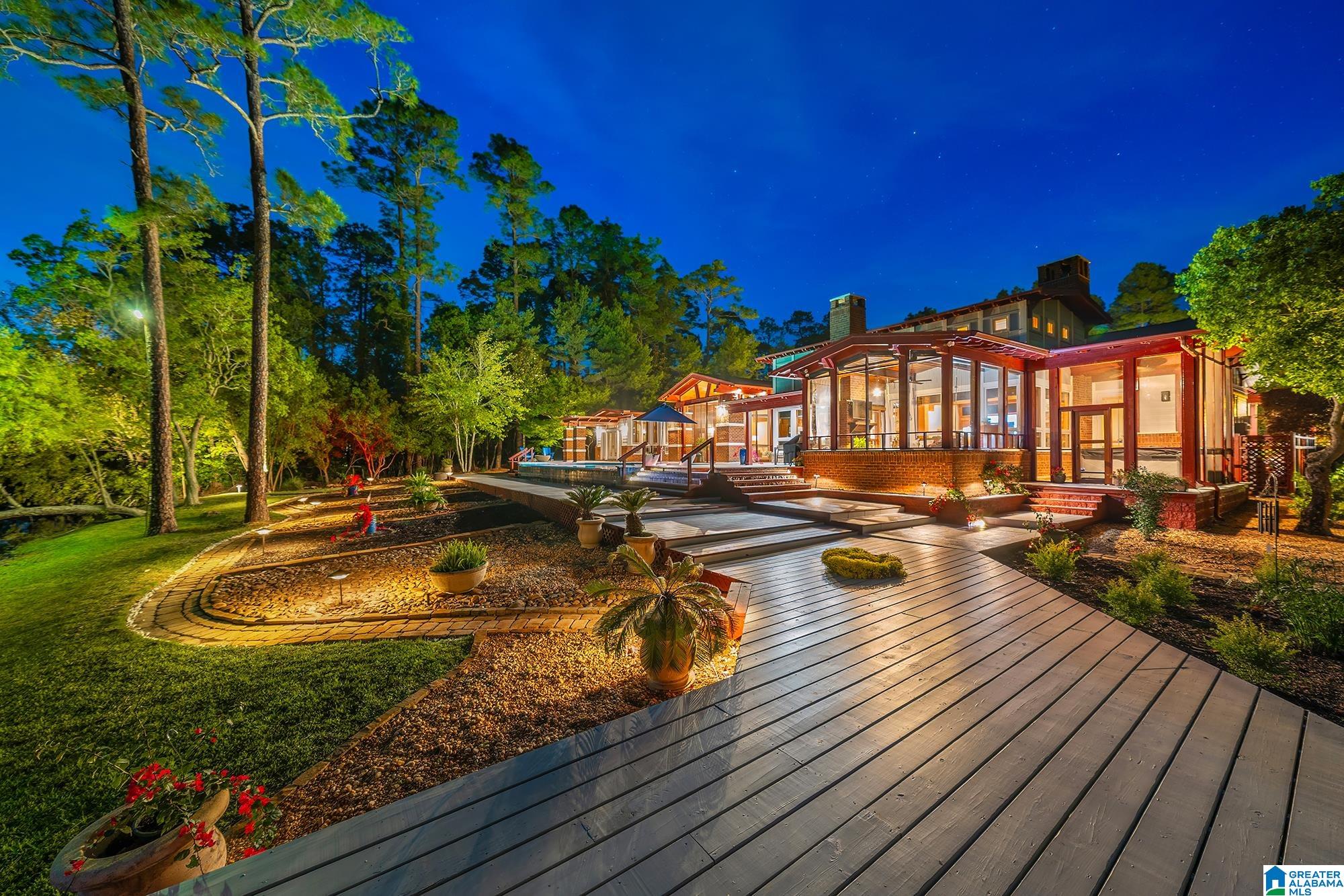 Illuminated glass-walled home beside a broad wooden deck and landscaped gardens at night.