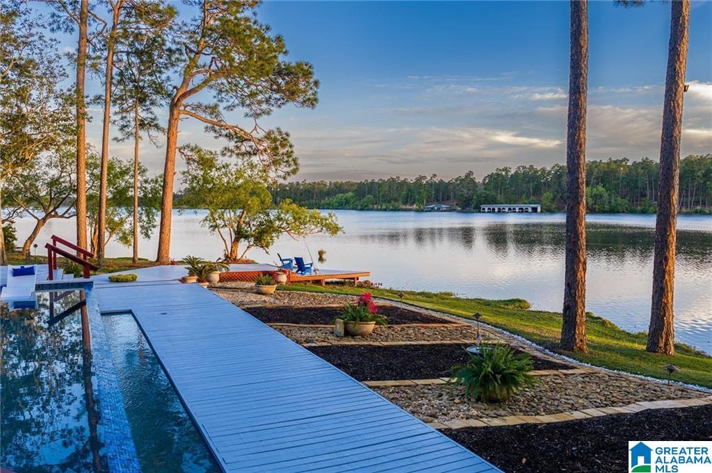 Lakeside pool and white walkway bordered by pine trees, patio chairs, and landscaped garden beds.