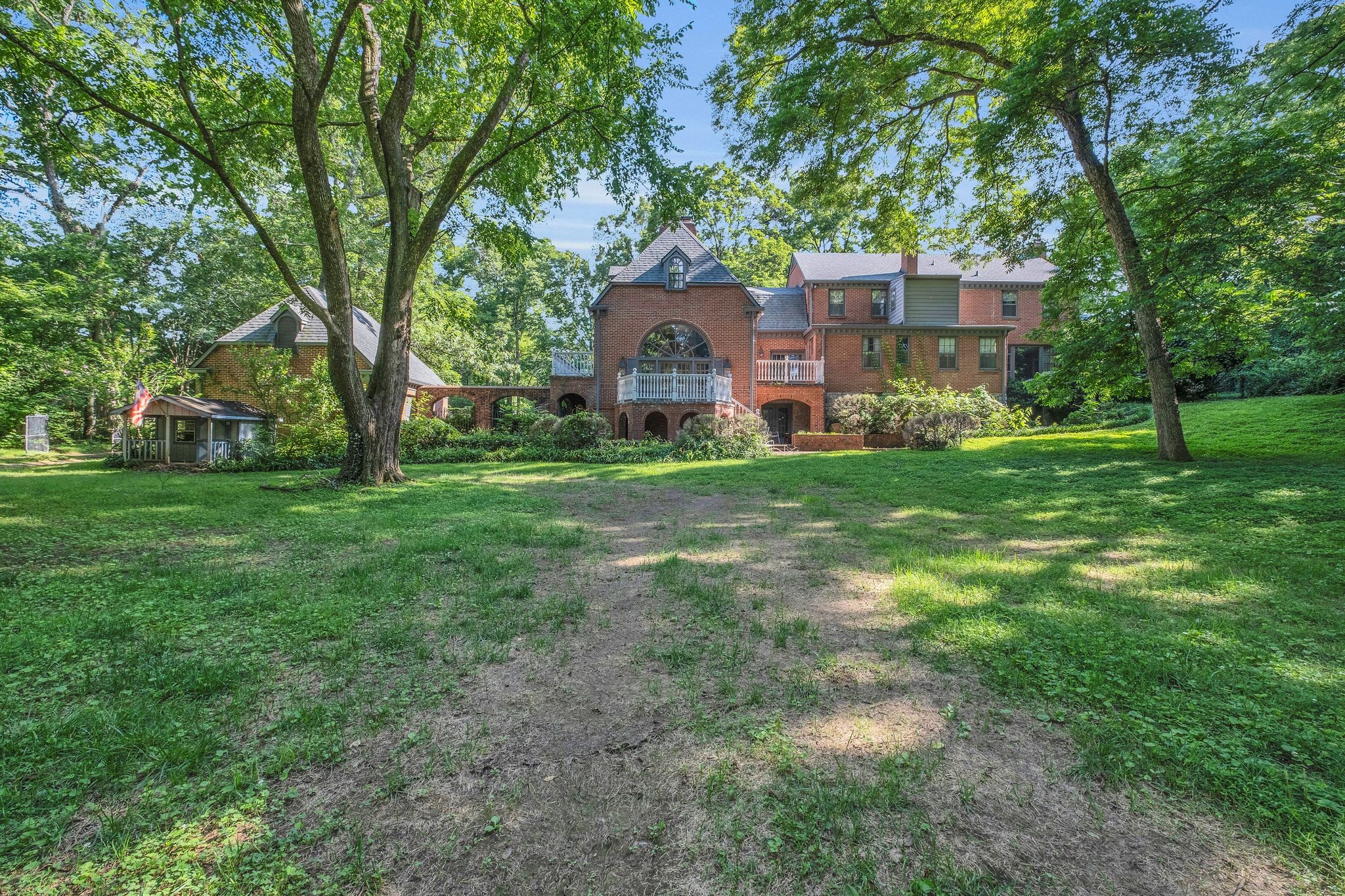 Red-brick home with balconies framed by mature trees on a spacious wooded lawn.