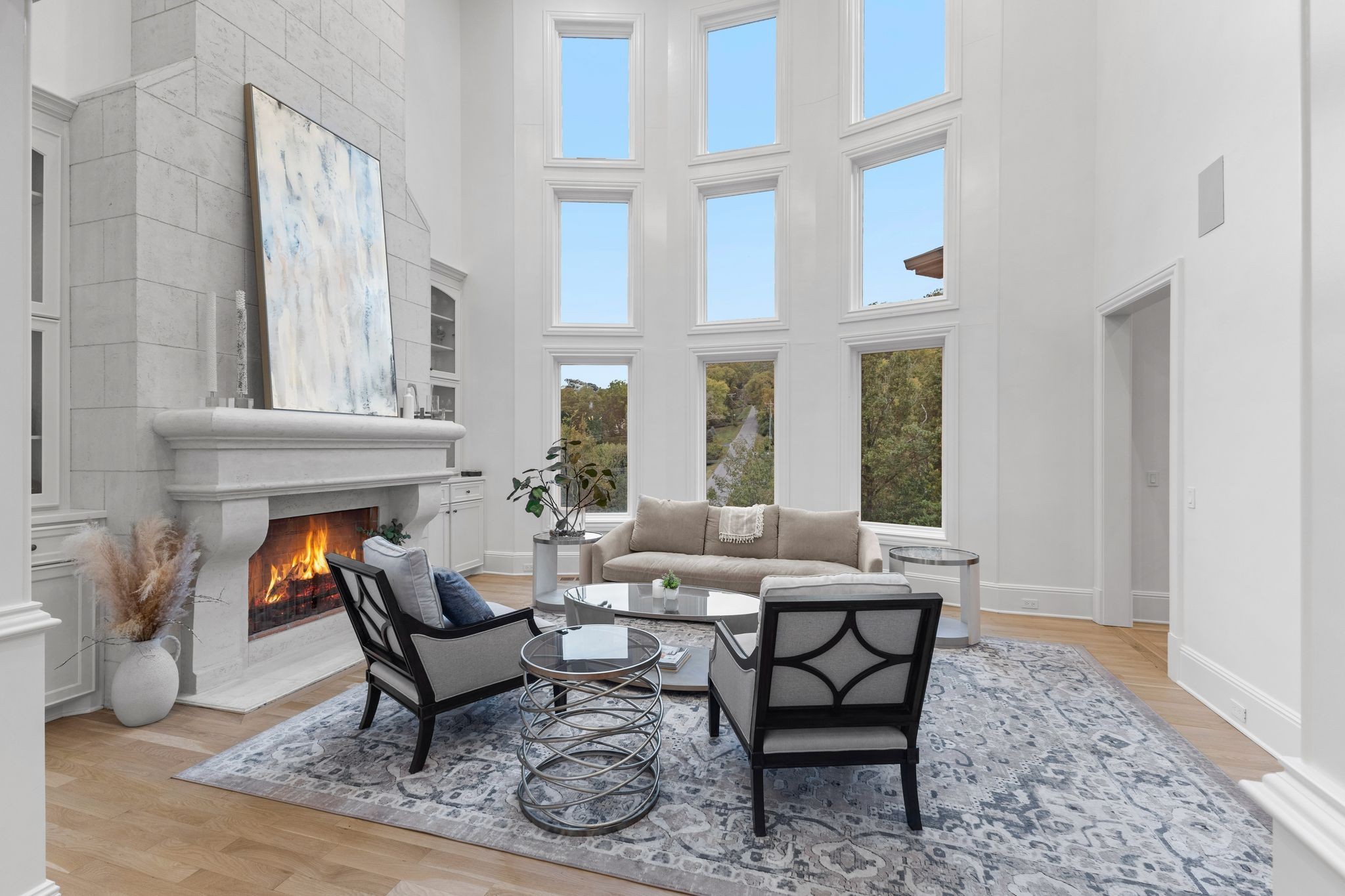 Staged living room with a tall stone fireplace, double-height windows, and neutral seating.