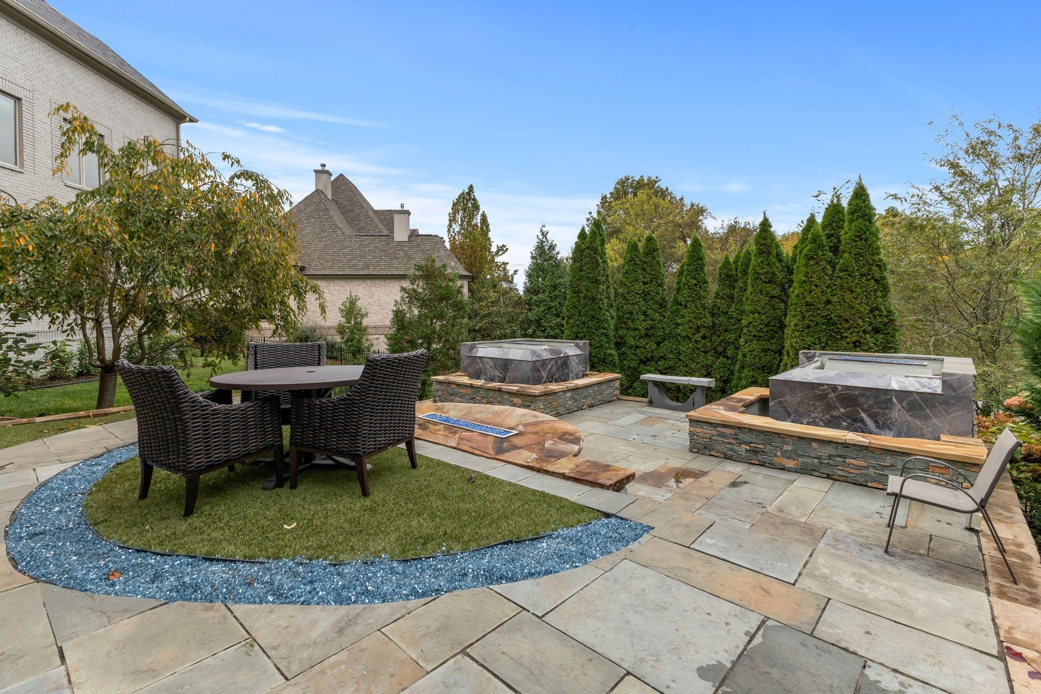 Backyard stone patio with woven dining chairs, raised stone water features, and tall evergreen hedges.