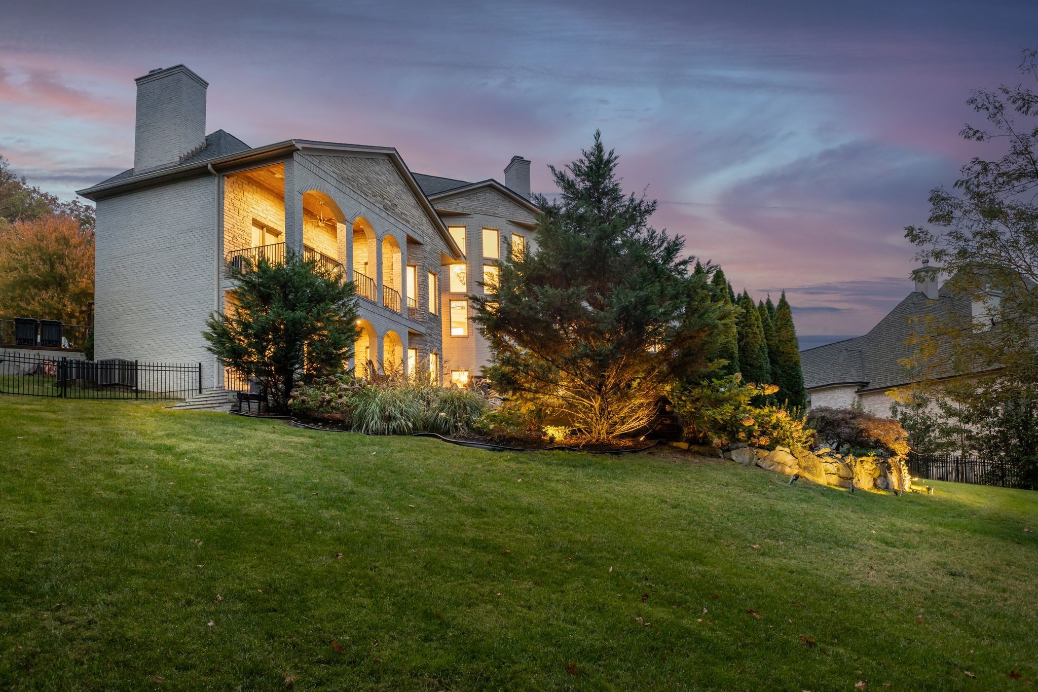 Two-story brick home with arched balconies and illuminated landscaping at dusk.