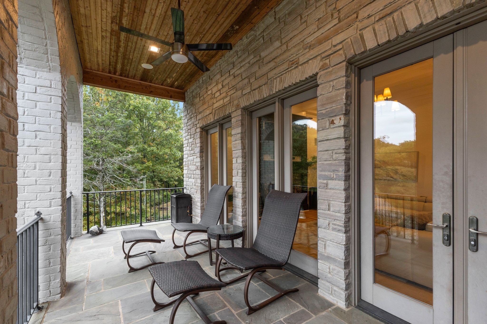 Covered stone patio with a vaulted wood ceiling, lounge chairs, and glass doors overlooking trees.
