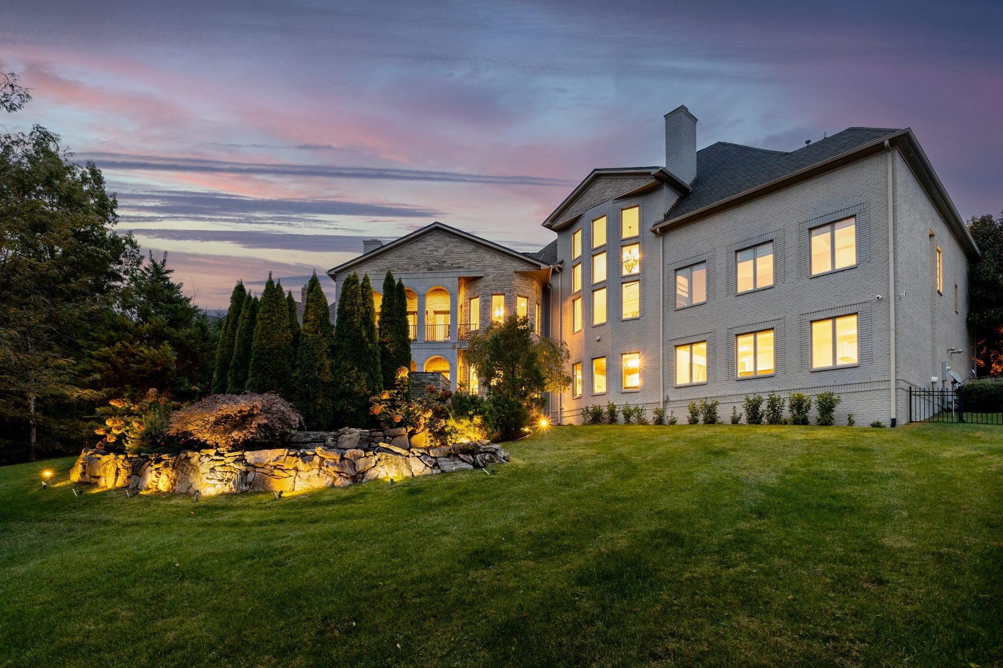 Large brick house at dusk with glowing windows, an arched stone entrance, and landscaped lawn.