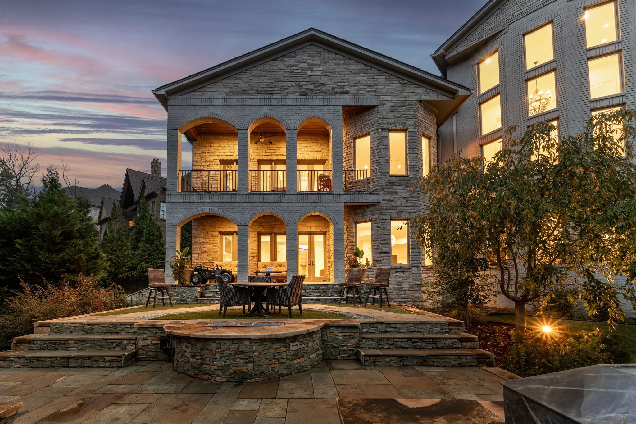 Stone house with arched porches and a tiered outdoor patio glowing at dusk.