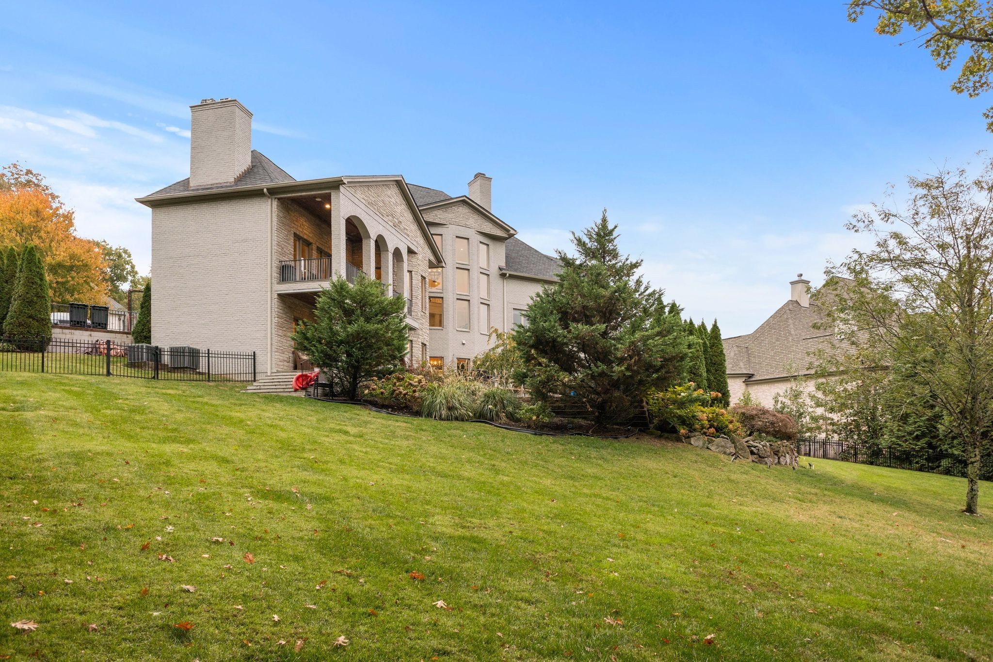 Rear of a light-brick house with an arched balcony, landscaped yard, and sloping lawn.